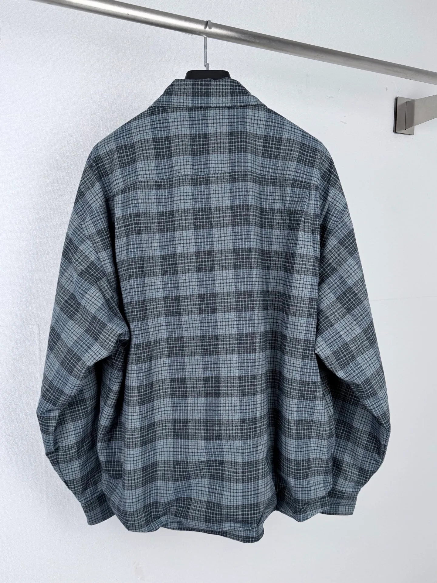 BALENCIAGA/balenciaga blue and gray plaid zipper pocket logo quilted jacket combines a three-layer structure of plaid woven fabric, filling cotton, and lining, and integrates plaid alignment technology and quilted garment production standards. There are multiple process difficulties in the whole process. The surface layer of the body is made of blue-gray Retro plaid fabric, and the cutting machine production strictly follows the global grid standards. The arrangement direction of the cutting pieces is fixed and cannot be changed at will. The horizontal and vertical lines of shoulder seams, side seams, and zipper placket joints must be consistent and coherent. The left chest patch pocket is cut independently, and the bag check pattern must be aligned with the front reference pattern. Slight deviations will destroy the visual integrity and directly increase fabric loss and cutting time. The most difficult part of the style is the three-layer composite sewing. The outer plaid fabric, filling cotton, and lining fabric have different physical properties, and the preshrinking processing parameters need to be adjusted separately. Pressure and temperature control are crucial during lamination operations. Once the parameters are out of balance, it can easily cause fabric stretch and distortion, plaid dislocation, and wrinkles and bulges in garments. At the same time, it is necessary to reasonably plan the quilting line to avoid the visual beauty of the grid pattern from being cut by the lines on the basis of firmly fixing the cotton layer and preventing cotton from running away. The placket adopts a centered zipper structure. It is difficult to sew the zipper tape and the quilted panels. It is required that the width of the left and right plackets be equal, the zipper routing is straight and smooth, and there is no lag in opening and closing. When the shirt lapel is filled with cotton, the thickness of the cotton needs to be controlled to prevent the collar from becoming bloated and unnaturally folded. The metal nameplate of the chest pocket is sewn at fixed points to avoid pulling the fabric during the operation and ensure that the plaid pattern is smooth and not deformed. The quilted material increases the overall thickness of the fabric. When sewing, you need to continuously adjust the presser foot pressure and stitch length to adapt to changes in sandwich thickness. The low-temperature and gentle ironing process is used for post-finishing to prevent the plaid from fading and the cotton layer from clumping due to high temperatures. From the precise alignment of cutting pieces, three-layer fabric pretreatment, composite quilting to garment finalization inspection, the processes are interlocking and the error tolerance rate is extremely low, testing the accuracy of plate making, equipment control and turning technology. Size:XS S M L